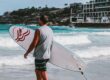 man walking towards body of water holding a surfboard